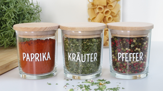 3 gefüllte Gewürzgläser mit Holzdeckel & mit weißen Etiketten mit der Aufschrift Paprika, Pfeffer, Kräuter