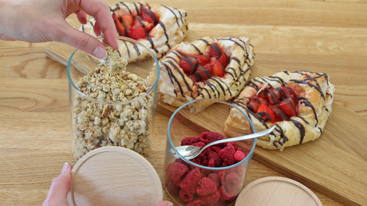 Eine Hand, die die sich Granola aus einem Vorratsglas nimmt. Daneben: Ein Glas mit gefriergetrockneten Himbeeren